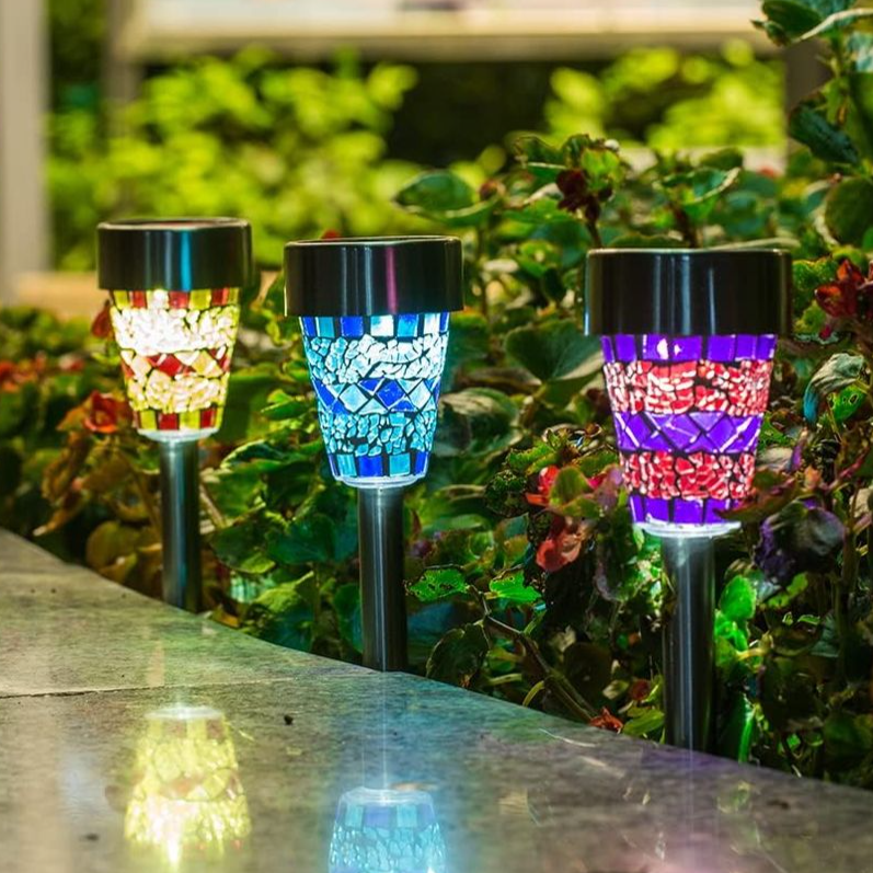 Three solar-powered garden lights with mosaic glass designs in red, blue, and multicolor hues are set along a garden path, illuminating nearby foliage.