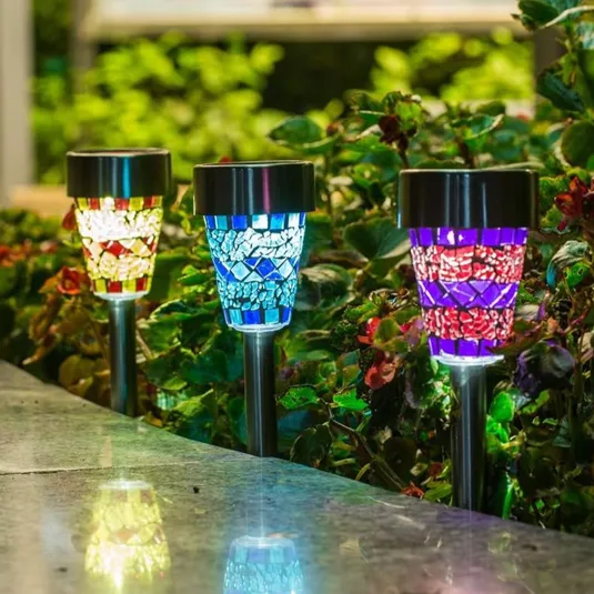 Three solar-powered garden lights with mosaic glass designs in red, blue, and multicolor hues are set along a garden path, illuminating nearby foliage.
