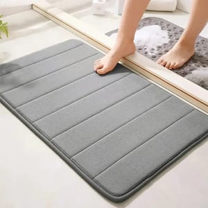 A grey bathroom mat with horizontal stripes and a dark border, beside a foot stepping out of a shower area.