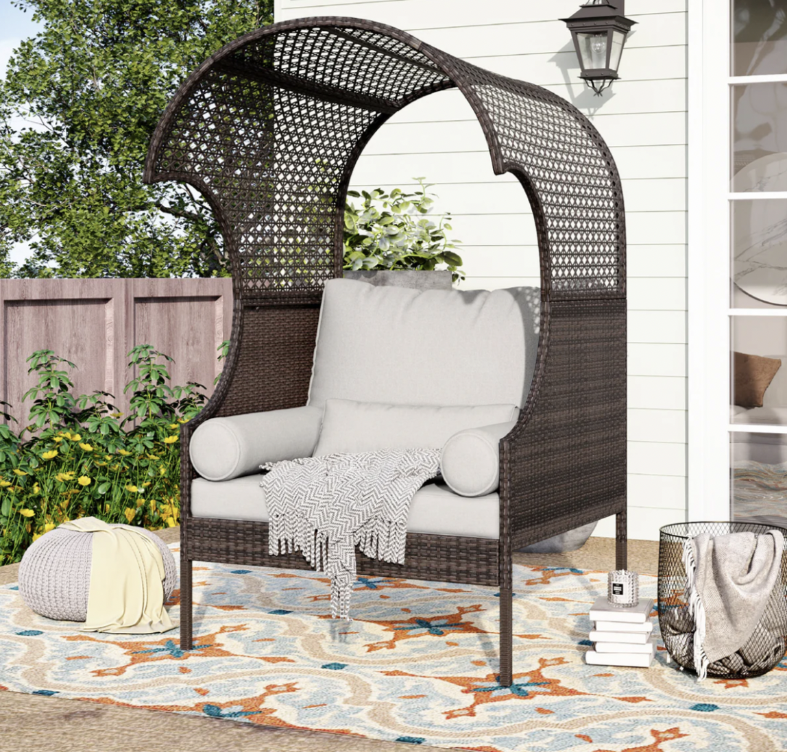 Wicker daybed with cushions and throw blankets, accompanied by a knitted pouf and outdoor rug.
