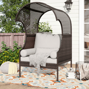 Wicker daybed with cushions and throw blankets, accompanied by a knitted pouf and outdoor rug.