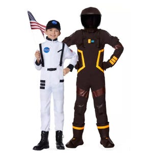Two children in astronaut-themed costumes, one in a white NASA suit holding a flag and the other in a brown flight suit with helmet.