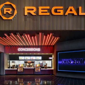 Regal cinema concessions area featuring a variety of snacks and drinks, with a popcorn station and digital menu displays. Bold signage and red lighting enhance the theater experience.