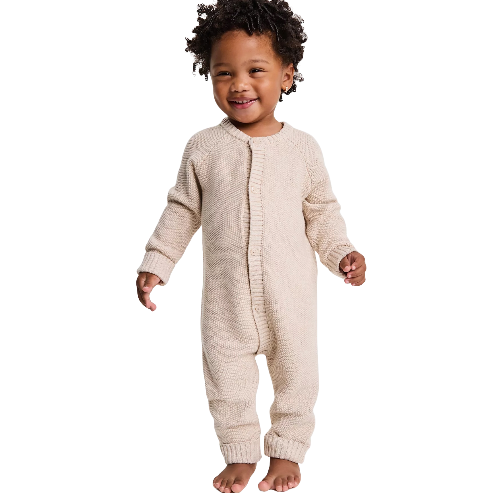 A toddler is wearing a beige, knitted, one-piece romper with buttons down the front, featuring long sleeves and closed feet.