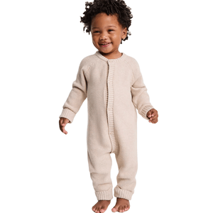 A toddler is wearing a beige, knitted, one-piece romper with buttons down the front, featuring long sleeves and closed feet.