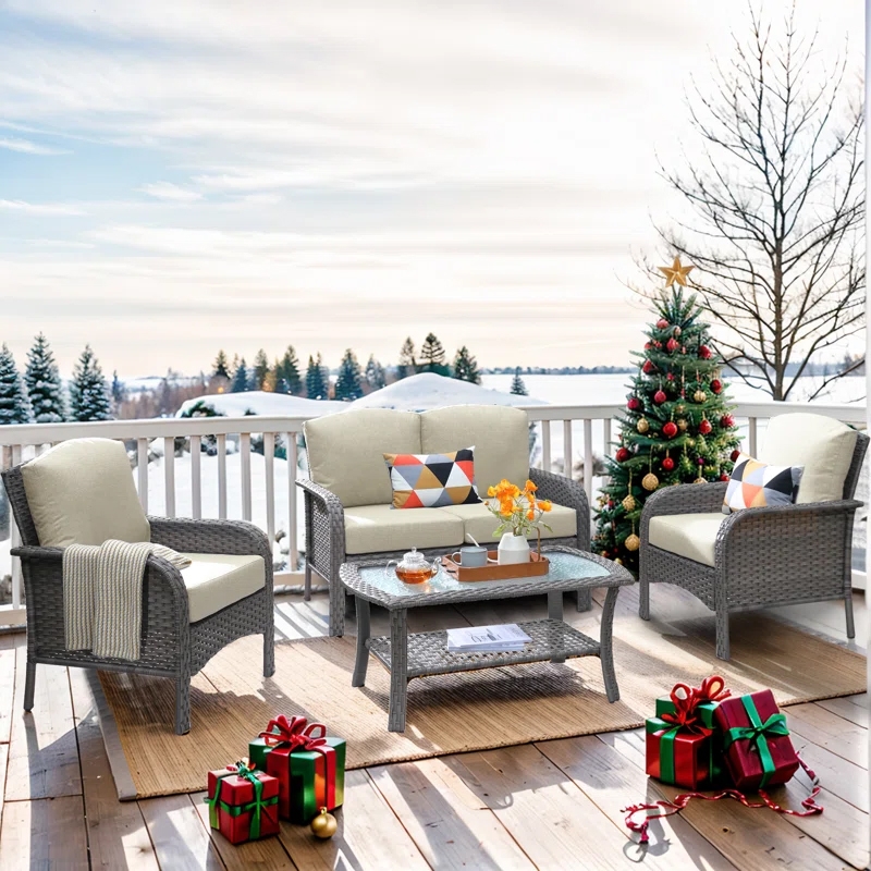 Outdoor wicker furniture set with cushions includes two chairs, a loveseat, and a glass-top coffee table, set on a rug. Christmas decor features a decorated tree and wrapped gifts. Snowy landscape and trees visible in the background.