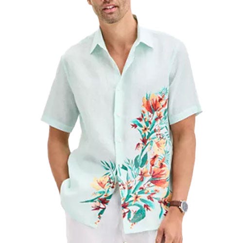 A man wears a light-colored short-sleeve button-up shirt with a floral design on one side, paired with white pants and a wristwatch.