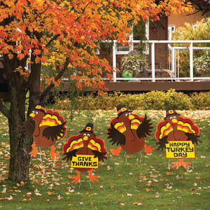 Four Thanksgiving-themed yard signs depict cartoon turkeys, two holding signs that say \“Give Thanks\“ and \“Happy Turkey Day,\“ set against a backdrop of a tree with autumn leaves.