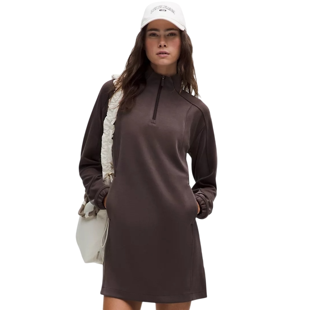 A woman is wearing a brown long-sleeve dress with a zip-up collar, a white bag slung over her shoulder, and a white cap.