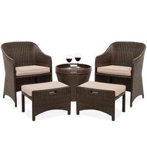 A five-piece patio set consisting of two wicker armchairs with beige cushions, two matching ottomans, and a round wicker table with a glass top and two wine glasses.