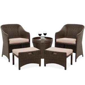 A five-piece patio set consisting of two wicker armchairs with beige cushions, two matching ottomans, and a round wicker table with a glass top and two wine glasses.