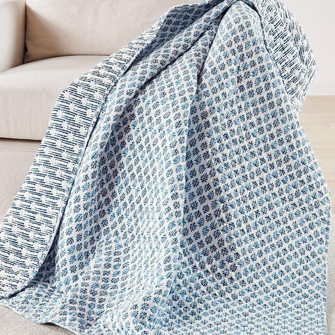 A quilted throw with a blue and white floral pattern on one side and a geometric design on the other, made of 100% cotton. Perfect for adding a cozy touch to a living space.