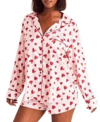 A woman is wearing a pajama set featuring a pink background with red heart patterns, consisting of a long-sleeve button-up top and matching shorts.