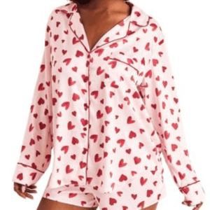 A woman is wearing a pajama set featuring a pink background with red heart patterns, consisting of a long-sleeve button-up top and matching shorts.