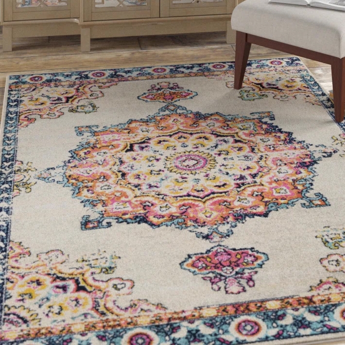 An ornate area rug with a central medallion design in vibrant colors including pink, orange, and blue, set against a light background. It's partially under a piece of furniture on a wooden floor.