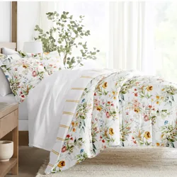 A bedding set with a floral design includes a duvet cover and matching pillowcases on a neatly made bed. The background features sheer curtains and a potted plant, creating a bright and airy bedroom atmosphere.