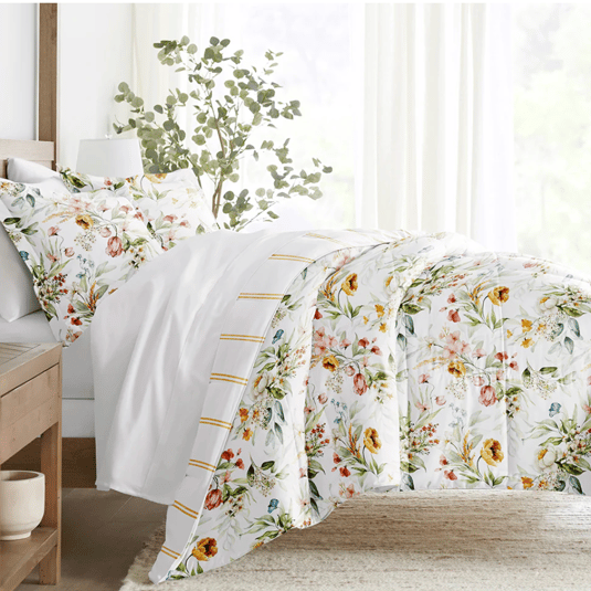 A bedding set with a floral design includes a duvet cover and matching pillowcases on a neatly made bed. The background features sheer curtains and a potted plant, creating a bright and airy bedroom atmosphere.