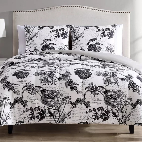A bed features a black and white floral and script-patterned comforter set with matching pillow shams against a light-colored, upholstered headboard.