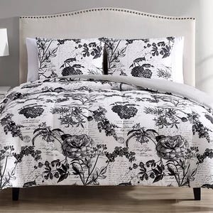 A bed features a black and white floral and script-patterned comforter set with matching pillow shams against a light-colored, upholstered headboard.