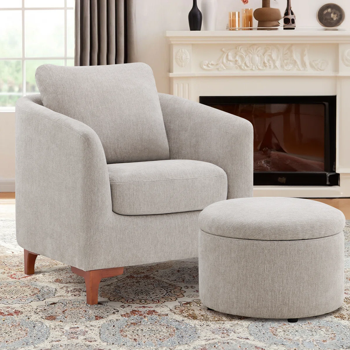 Light gray upholstered armchair with a curved design, accompanied by a matching round ottoman. The setup is in a cozy room featuring a patterned rug, curtains, and an ornate fireplace with decorative items.