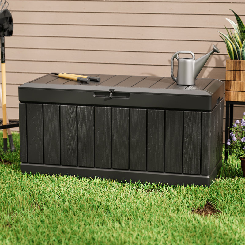 A black outdoor storage bench with a shovel and trowel on top, next to a metal watering can.