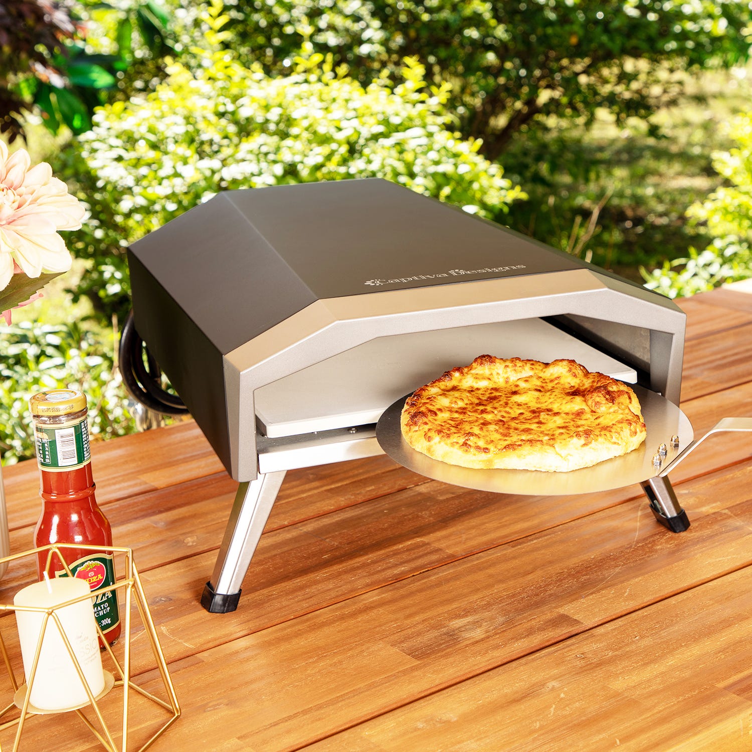 A portable outdoor pizza oven with a freshly baked pizza on a peel and a bottle of seasoning next to it.