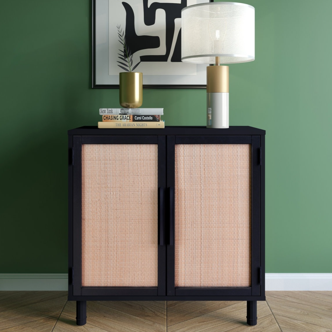 A black cabinet with woven rattan doors is topped with a gold and white table lamp, a gold vase, and three stacked books against a green wall with abstract art.