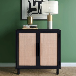 A black cabinet with woven rattan doors is topped with a gold and white table lamp, a gold vase, and three stacked books against a green wall with abstract art.