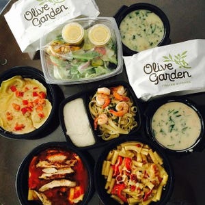 A variety of Olive Garden dishes including pasta with shrimp, ravioli, chicken with sauce, a container of soup, salad with dressing packets, and a side of sauce.