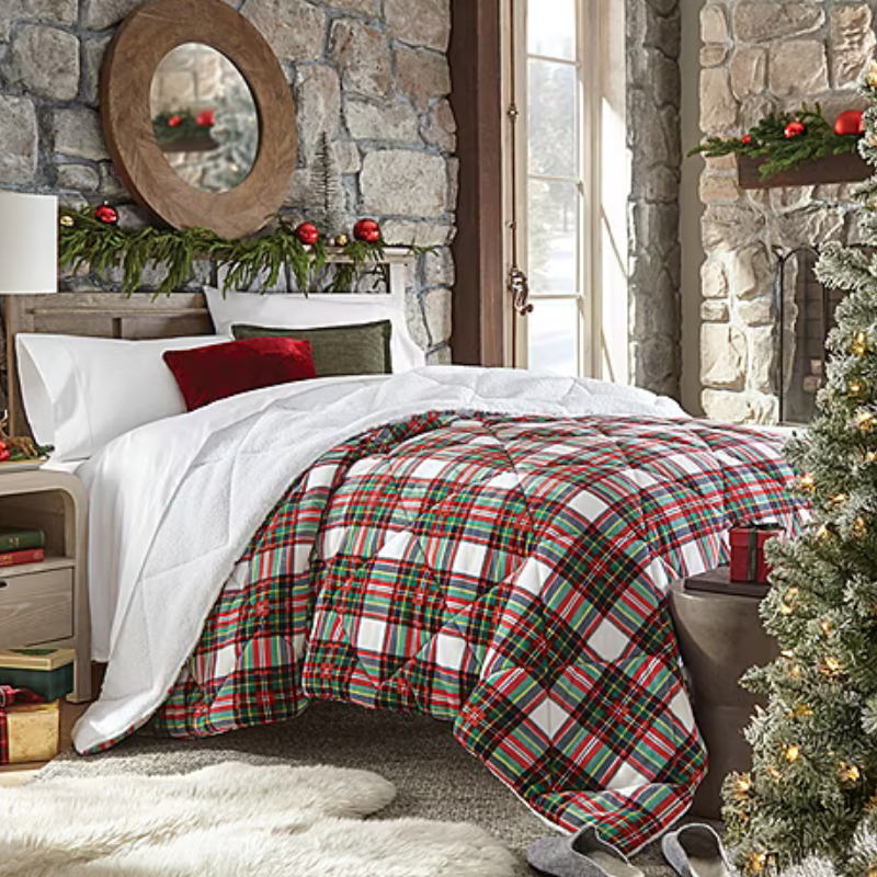 A festive bedroom features a plaid comforter with red, green, and white accents, flanked by decorative holiday elements like a wreath above the headboard and a Christmas tree beside the bed. The room has stone walls, a circular mirror, and cozy textiles.
