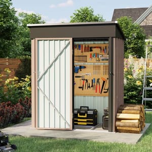 A small metal storage shed with a sliding door, featuring a tool organizer on the interior wall with various hand tools and power tools. Firewood is stacked outside on a concrete slab, surrounded by a garden with flowers and trees.