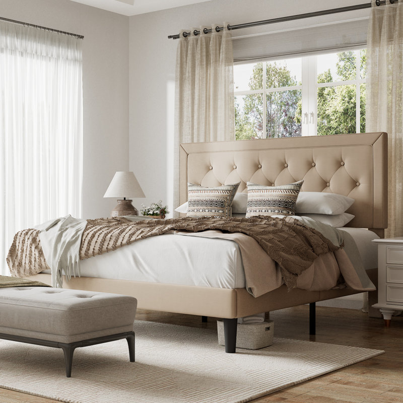 A beige, upholstered, tufted queen platform bed is featured with a padded headboard and short, sturdy legs. It has decorative pillows, neutral bedding, and is complemented by a cozy throw, set in a bright, airy bedroom with beige curtains.