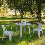 A small round table and two metal chairs are set up on grass under trees, with a plate of fruit and a cup on the table, creating a serene outdoor scene.