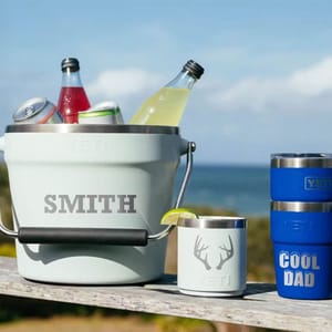 A YETI bucket labeled \“SMITH\“ holds beverages. Nearby are a YETI mug with a deer design and two stacked blue YETI tumblers labeled \“COOL DAD.\“ The scene is set outdoors with an ocean backdrop.