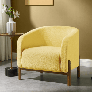 A cozy yellow armchair with wooden legs is next to a small side table holding a white vase with flowers and accompanied by a black and gold floor lamp. The setting is in a modern, warmly lit room with an abstract painting on the wall.