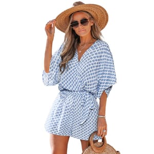 Woman wearing a blue and white patterned romper with a cinched waist, accessorized with a wide-brimmed straw hat, sunglasses, and holding a woven bag.