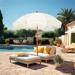 A 7' white patio umbrella with a fringed edge stands on a poolside area, shading two cushioned lounge chairs with yellow pillows on a light stone patio.