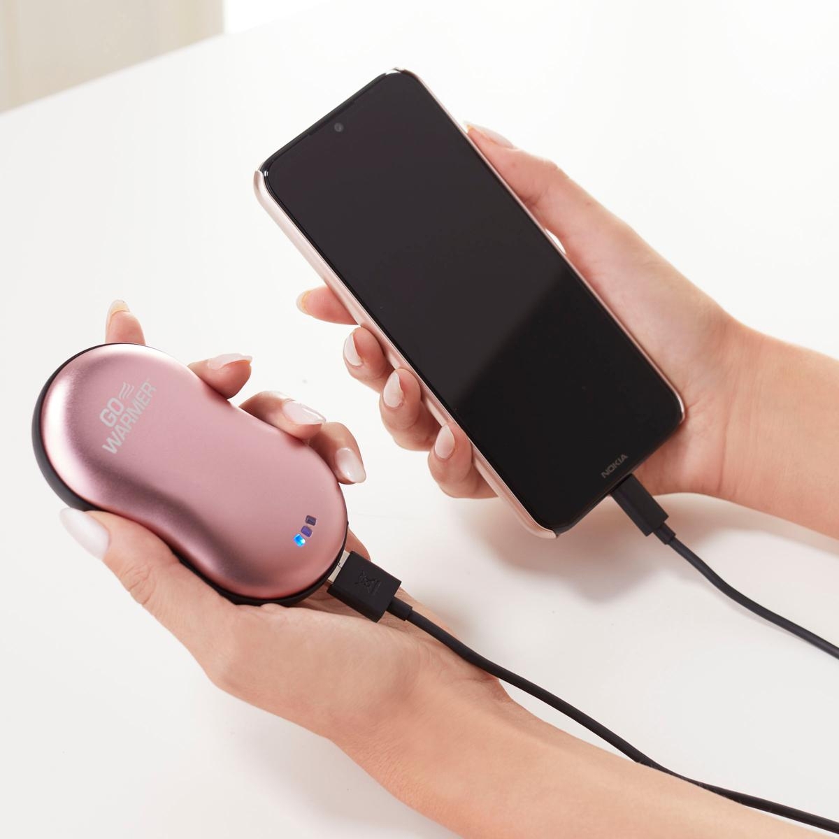 Two hands are holding a smartphone connected to a pink, palm-sized portable power bank that doubles as a hand warmer, with a blue LED indicator visible on the device.