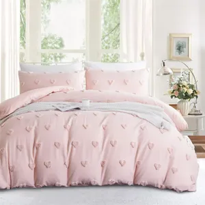 A pink bedding set with heart-shaped texture, including a comforter, two pillowcases, and a knitted throw blanket, is styled on a bed. A side table holds a lamp and a vase of flowers, situated by large windows with a view of greenery.