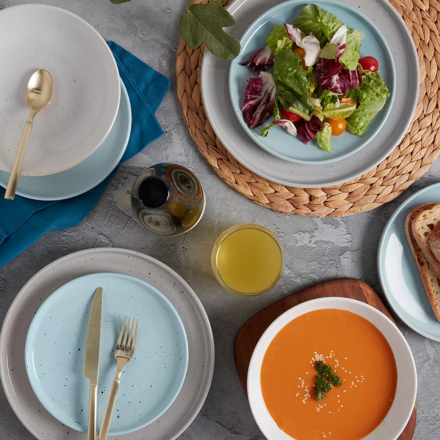 A table setting features a mixed greens salad with cherry tomatoes, creamy soup, and toast. There's a glass of orange juice, a small bottle possibly for dressing, and gold cutlery on speckled plates with a blue napkin.