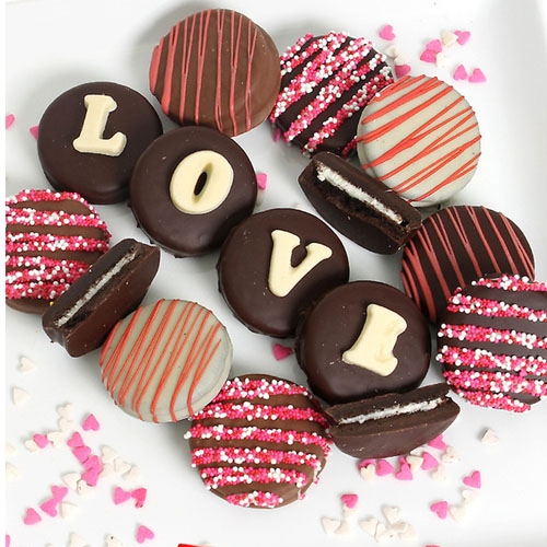 Assorted chocolate-covered cookies decorated with red and pink sprinkles, stripes, and white letters spelling \“LOVE,\“ surrounded by small heart-shaped sprinkles.