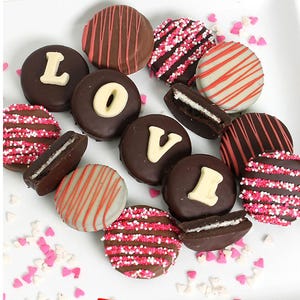 Assorted chocolate-covered cookies decorated with red and pink sprinkles, stripes, and white letters spelling \“LOVE,\“ surrounded by small heart-shaped sprinkles.