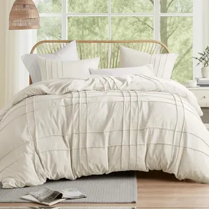 A neatly made bed with a textured cream-colored duvet and multiple pillows, set against a wooden headboard. A side table with a plant and a mug, plus an open magazine on a rug, complete the cozy bedroom scene.