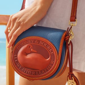 Dooney & Bourke All-Weather Duck Bag features a blue pebbled leather body with brown leather accents, a large embossed duck logo, gold hardware, and a crossbody strap.