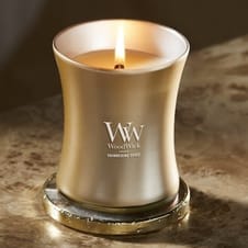 A gold-colored WoodWick candle labeled \“Shimmering Spice\“ with a visible flame sits on a round coaster.