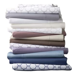 A stack of folded bed sheets in various colors and patterns, including shades of white, gray, blue, brown, and lavender, with some featuring geometric designs.