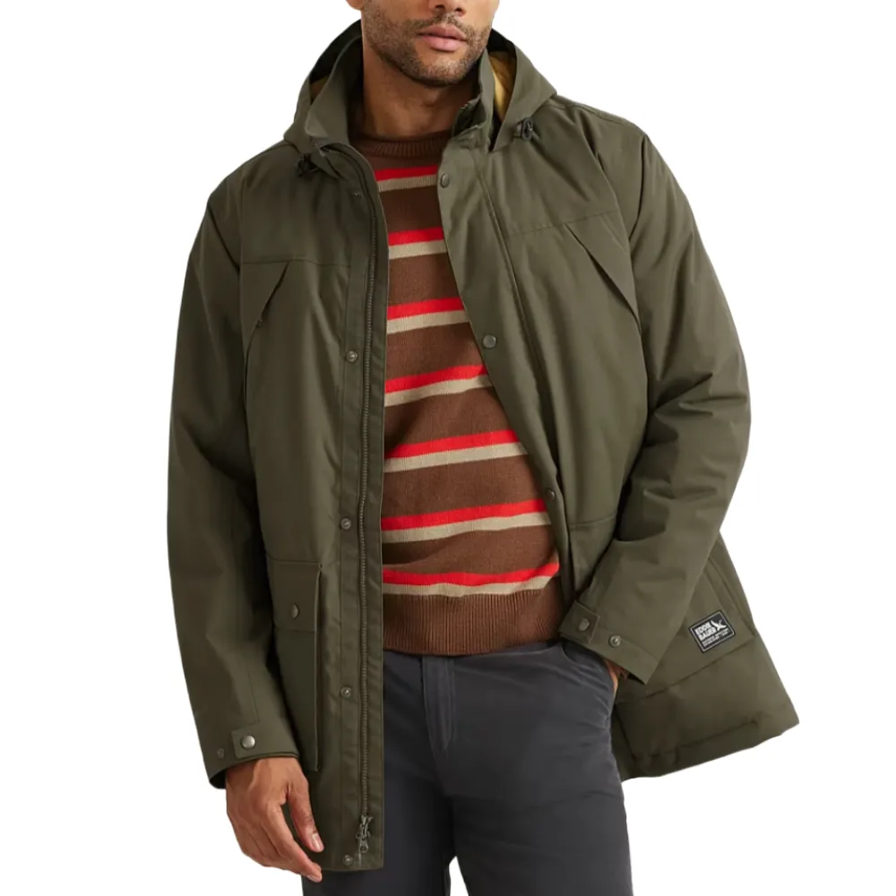 A man wears an olive-green hooded jacket over a brown sweater with red and beige stripes, paired with dark pants.