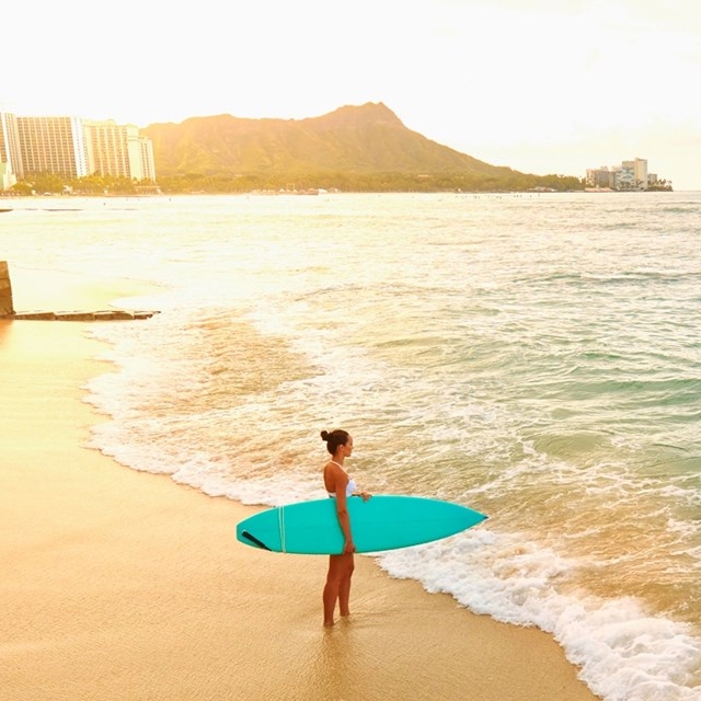 A person stands on a beach with a turquoise surfboard, gazing at the ocean. In the background, a cityscape and a recognizable mountain are visible.