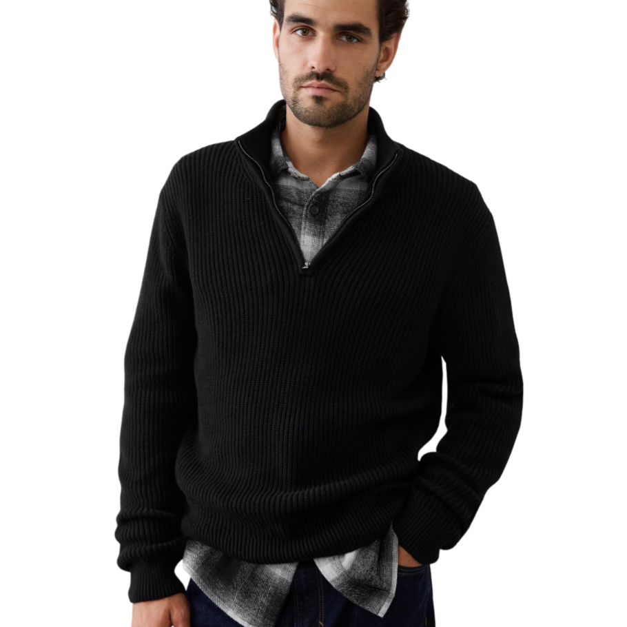 A man wears a black ribbed zip-up sweater over a black and white plaid shirt, paired with dark jeans.