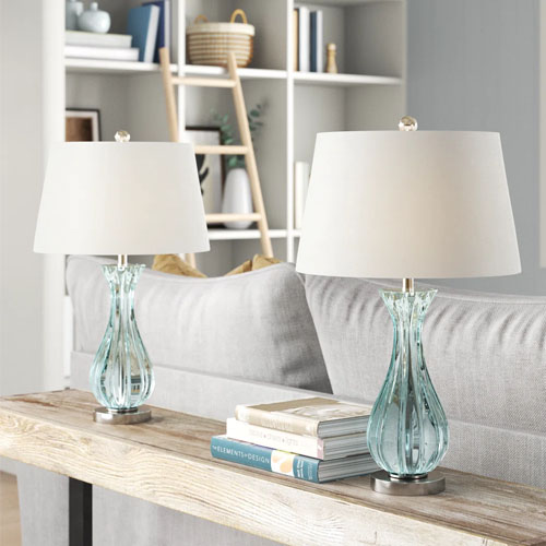 Two elegant table lamps with clear glass bases and white shades are placed on a console table behind a sofa.